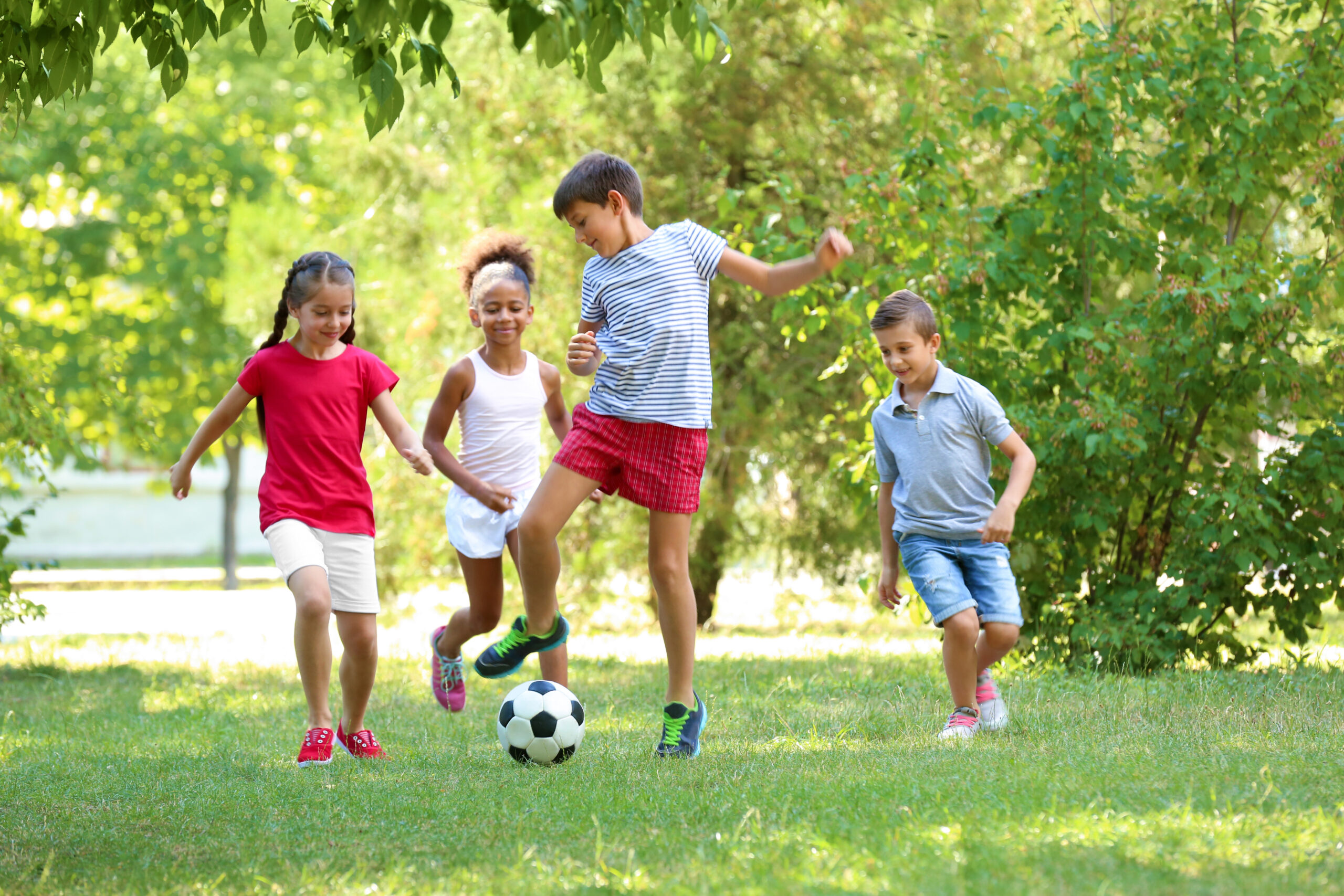 Four diverse children play soccer on a sunny, green lawn surrounded by lush summer foliage.