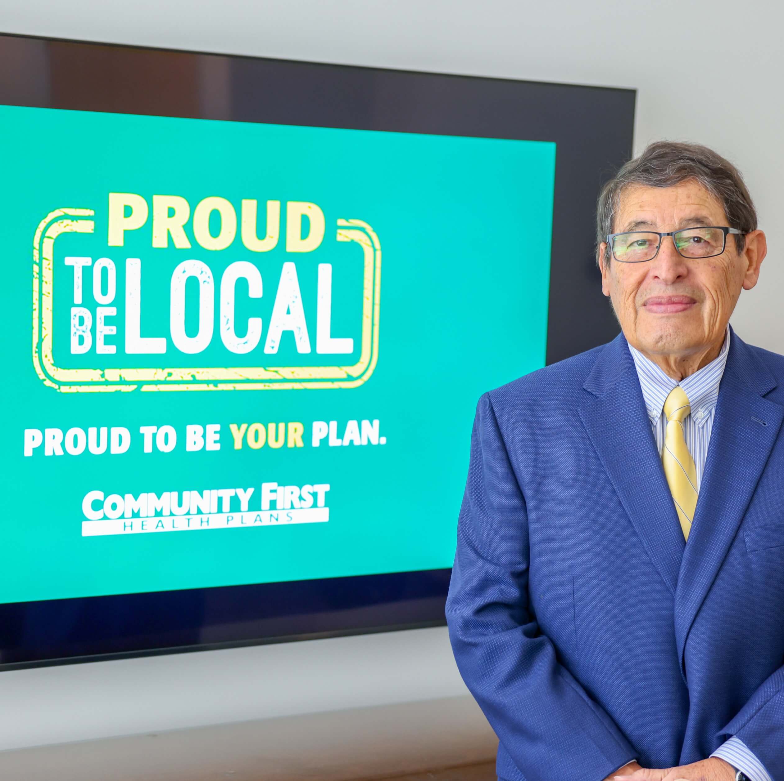 George B. Hernández Jr in a blue suit stands in front of a screen displaying "Proud to be local. Proud to be your plan."