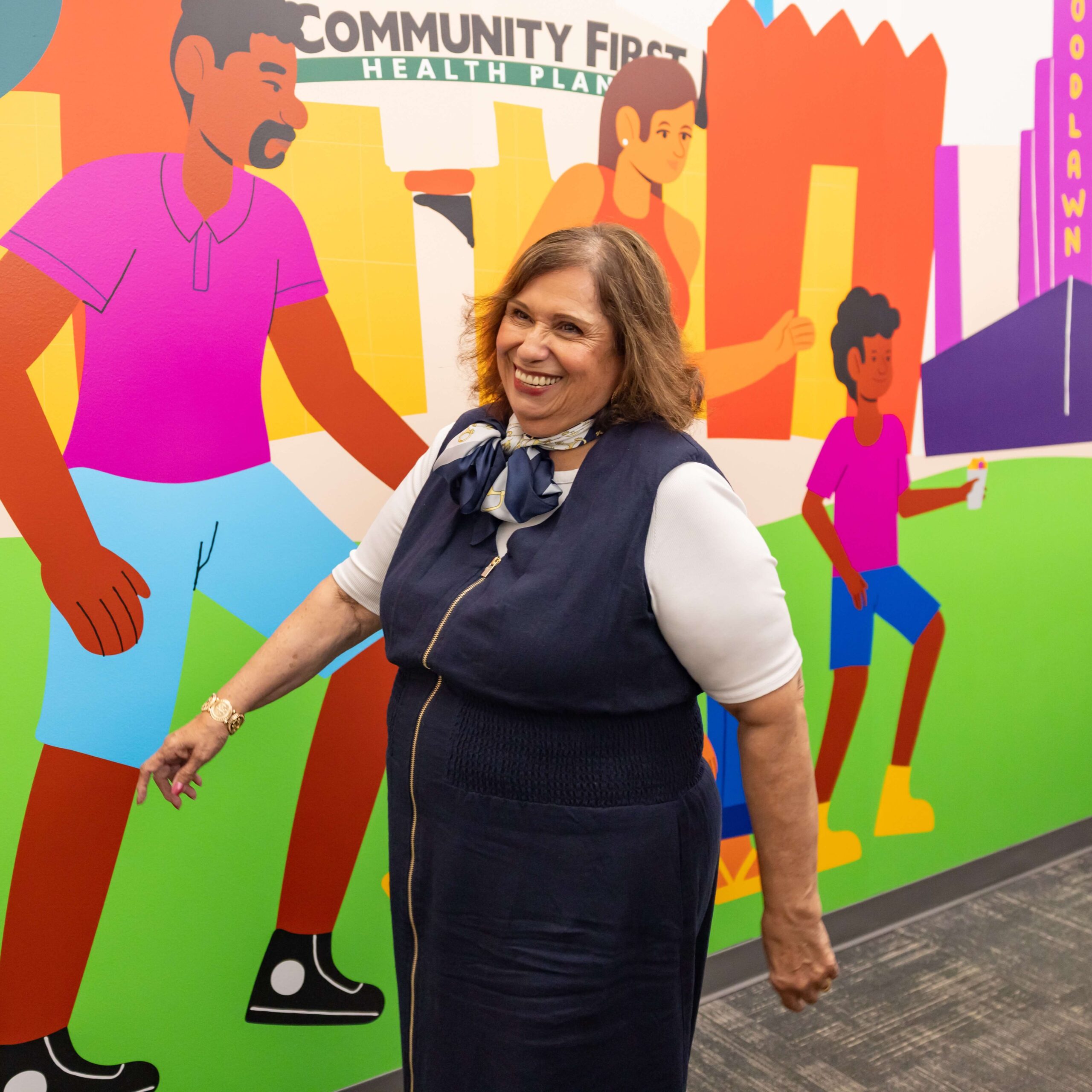 Mary Helen Gonzalez smiling in front of a colorful mural featuring people and the text "Community First Health Plan".