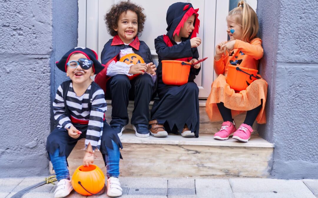 Four children in Halloween costumes, including a pirate, a vampire, and a pumpkin, sit on steps holding candy buckets.