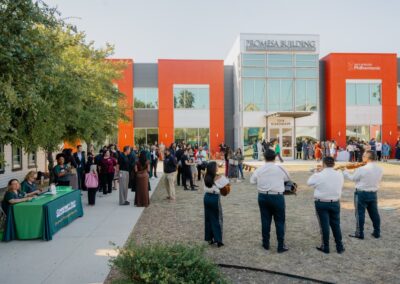 A diverse group gathers outdoors for an event in front of the modern Promesa Building, featuring a mariachi band playing on the lawn.