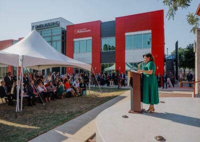 Judy Razo speaks at a podium outdoors during the 30 over 30 Celebration in front of a modern red and grey La Promesa building.