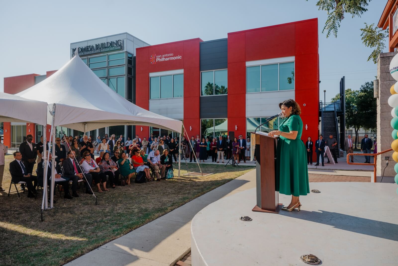 Judy Razo speaks at a podium outdoors during the 30 over 30 Celebration in front of a modern red and grey La Promesa building.