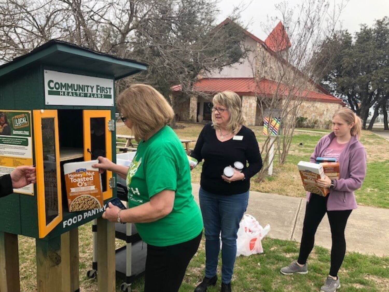 Three women stock a green outdoor Community First Health Plans food pantry with food outside a stone building.