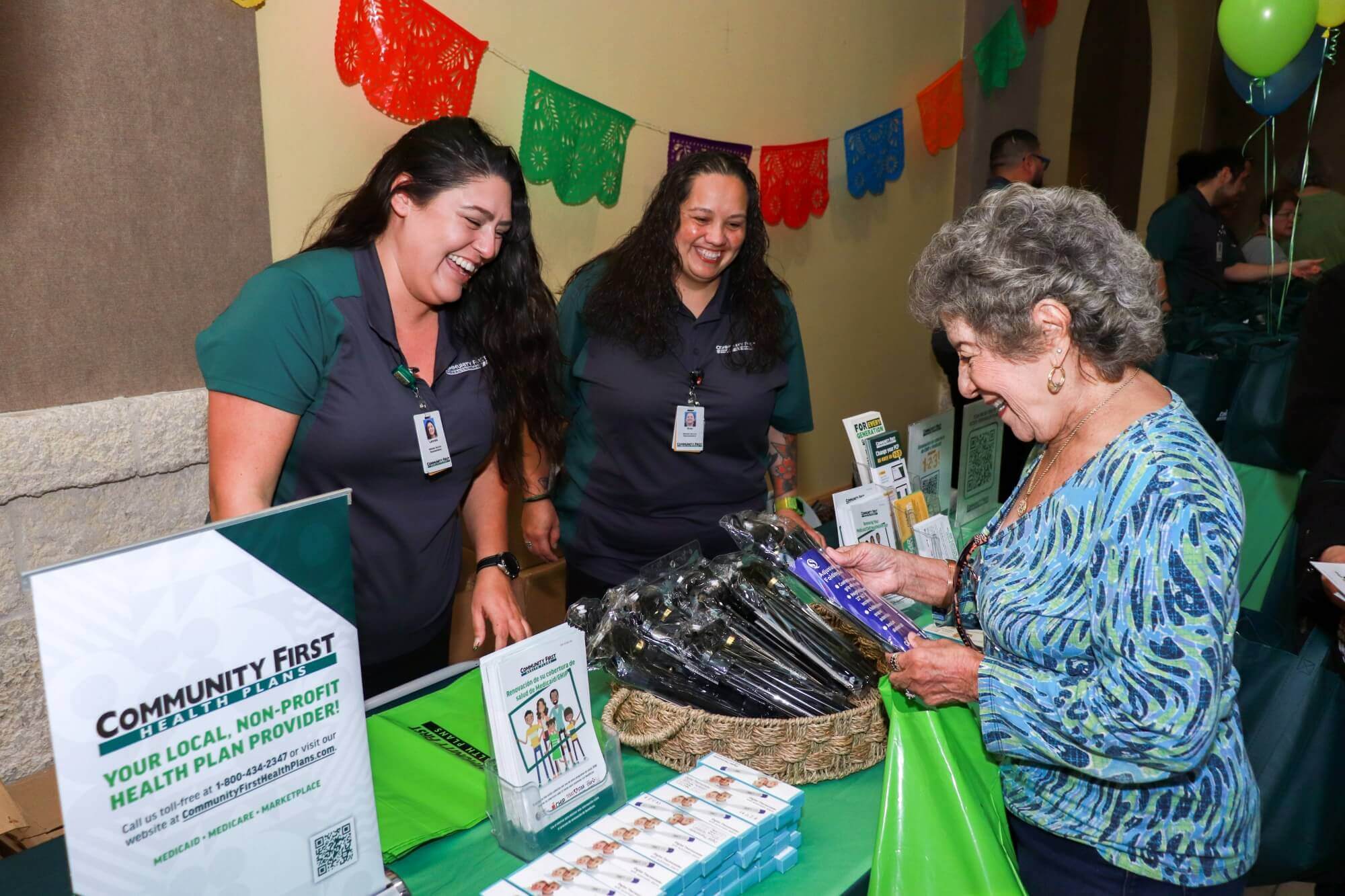 Two smiling staff members in matching shirts offer promotional items to an older woman at a Community First Health Plans information booth decorated with colorful papel picado banners.