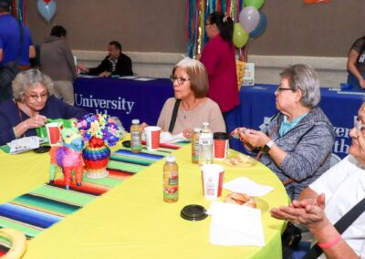 Four older women sit at a bright yellow table decorated with a colorful serape runner, piñata, and refreshments, with event booths in the background.