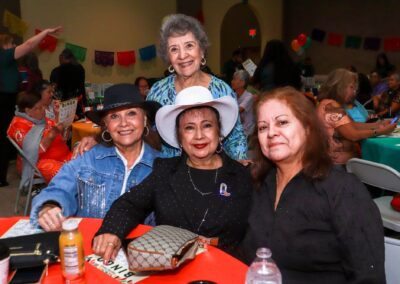 Four smiling Latina women pose closely together at a brightly decorated indoor event, possibly a Bingo game, with other attendees visible in the background.