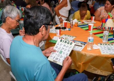 An older man in a blue shirt focuses on a "Healthy Habits BINGO" card while seated at an orange-covered table with several other attendees in a crowded indoor setting.