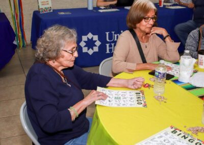 Two older women sit at a bright yellow table playing bingo with small markers during a community event sponsored by a health organization visible in the background.