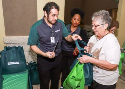A male staff member in a green and gray polo shirt hands a reusable tote bag to an older woman while a female colleague stands nearby at an indoor event with branded bags on a table.