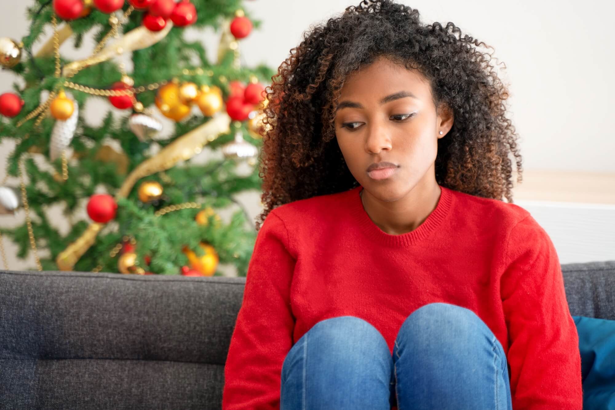 A young Black woman in a red sweater sits sadly on a couch with a blurred, decorated Christmas tree in the background.