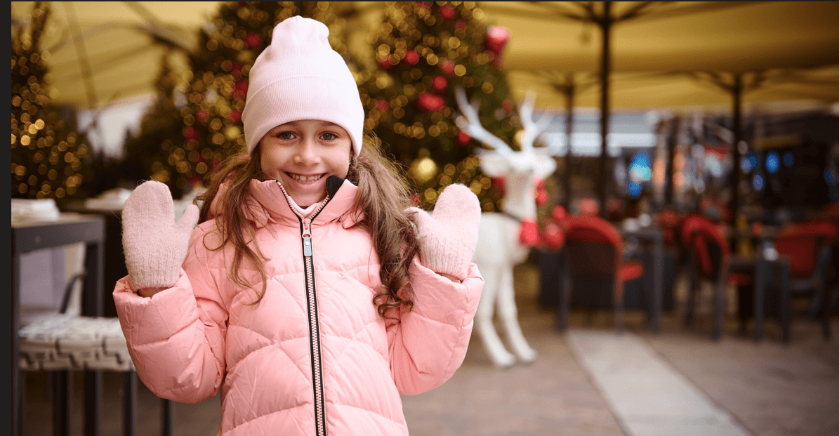 Smiling young girl in a pink winter coat, hat, and mittens stands outdoors near blurred Christmas decorations including lights and a white reindeer figure.