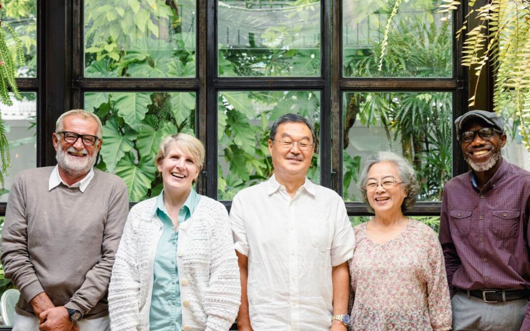 Five diverse, smiling senior adults stand together indoors in front of a large window showcasing dense green foliage.