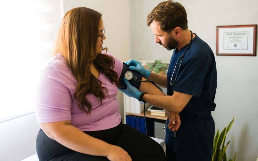 Bearded male nurse in blue scrubs checks the blood pressure of a female patient wearing a lavender shirt in a brightly lit medical office.