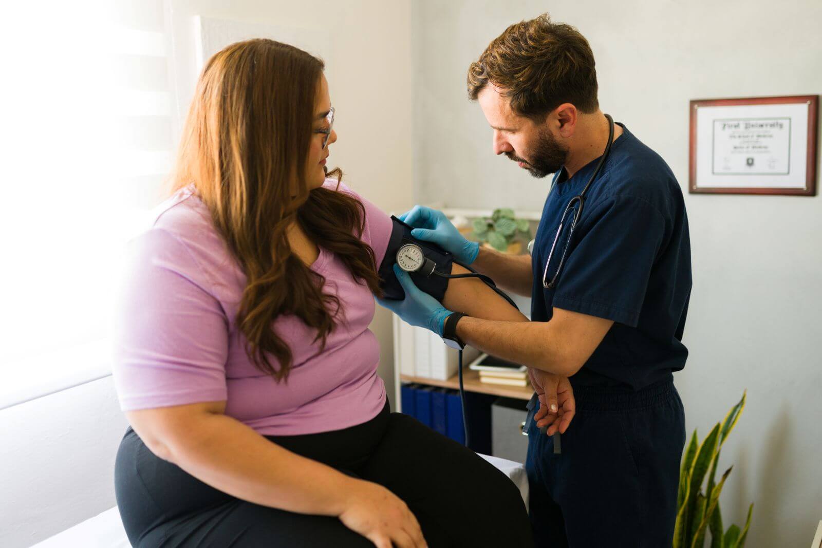 Bearded male nurse in blue scrubs checks the blood pressure of a female patient wearing a lavender shirt in a brightly lit medical office.