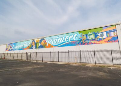 Large, colorful outdoor mural reading "Comunidad Primero" featuring portraits, nature, and neighborhood scenes, set against a white building facade above a black metal fence and empty parking lot.