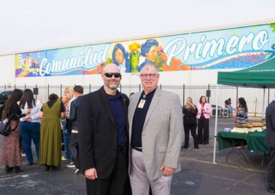 Two men pose for a photo outdoors in front of a large mural reading "Comunidad Primero" at an event with a crowd gathered nearby.