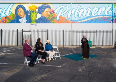 A woman speaks at a podium during an outdoor event in front of a large mural featuring portraits and the word "Primero."