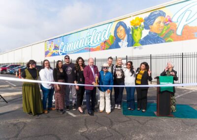 A diverse group of people participates in a ribbon-cutting ceremony in front of a large outdoor mural featuring the word "Comunidad Primero" and portraits of historical figures.