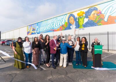 A diverse group celebrates a ribbon-cutting ceremony in front of a large white wall featuring a vibrant mural that reads "Comunidad Primero" with portraits of historical figures.