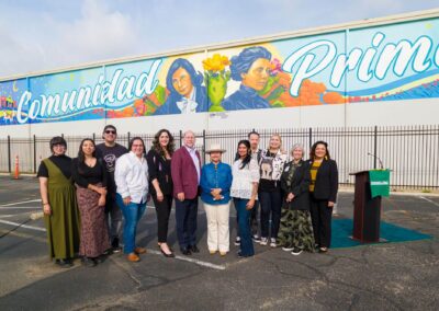 A diverse group celebrates a ribbon-cutting ceremony in front of a large white wall featuring a vibrant mural that reads "Comunidad" with portraits of historical figures.