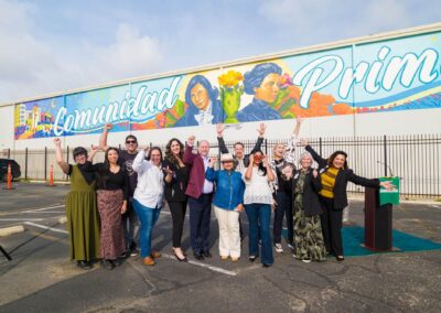 A diverse group celebrates a ribbon-cutting ceremony in front of a large white wall featuring a vibrant mural that reads "Comunidad" with portraits of historical figures.