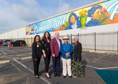 Five smiling people stand outdoors in front of a large, colorful mural featuring the word "Comunidad" and historical figures.