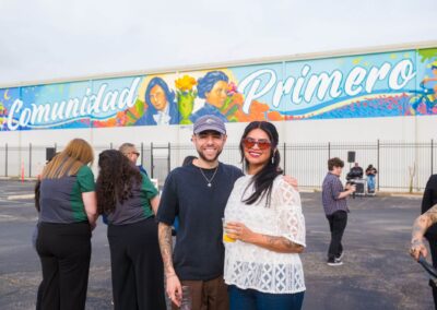 Two smiling people stands outdoors in front of a vibrant mural reading "Comunidad Primero," with several other people gathered in the background.