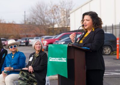 Professional woman speaking at an outdoor podium featuring a green Community First Health Plans banner, with attendees seated behind her in front of a parking lot.