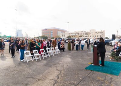 A woman speaks at a podium outdoors to a crowd gathered in a paved lot with white folding chairs, backed by parked cars and city buildings under an overcast sky.