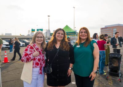 Three smiling women pose outdoors in a busy event setting with cars, a tent, and highway signs visible in the background.