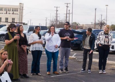 A diverse group of people stands in an outdoor parking lot in front of several parked cars, with one woman crouching in the foreground taking a photo with her iPhone.