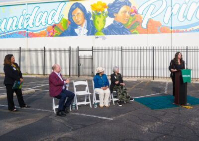 A group of people attend an outdoor presentation in front of a large mural featuring portraits and the Spanish word "Comunidad Primero" above a black metal fence.