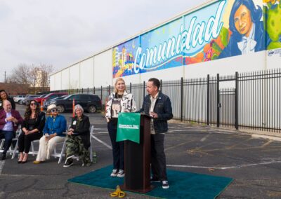 Two people stand at a podium outdoors in front of an audience, with a large colorful mural featuring the word "Comunidad" on the building wall behind them.