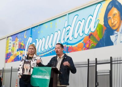 A man speaks at a podium draped with a Community First Health Plans banner while a woman stands beside him in front of a colorful mural featuring the word "Comunidad" and a portrait.