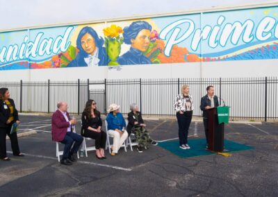 A group of people attend an outdoor speaking event in a paved lot beneath a colorful mural featuring portraits and the word "Comunidad Primero."