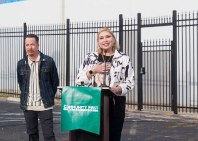 A woman in a patterned jacket speaks at a podium displaying a "Community First Health Plans" banner, while a man in a denim jacket stands to her left outdoors against a black metal fence.
