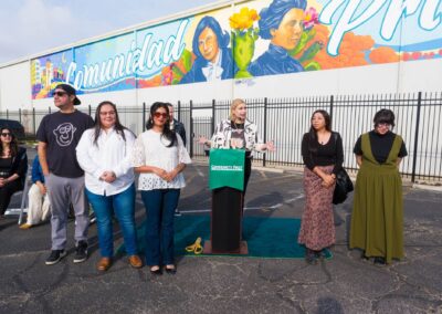 A group of diverse individuals stands outdoors for a press conference in front of a large mural displaying the word "Comunidad Primero" and portraits of historical women.
