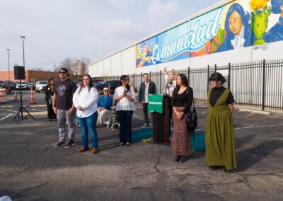 A group of diverse individuals stands outdoors for a press conference in front of a large mural displaying the word "Comunidad Primero" and portraits of historical women.