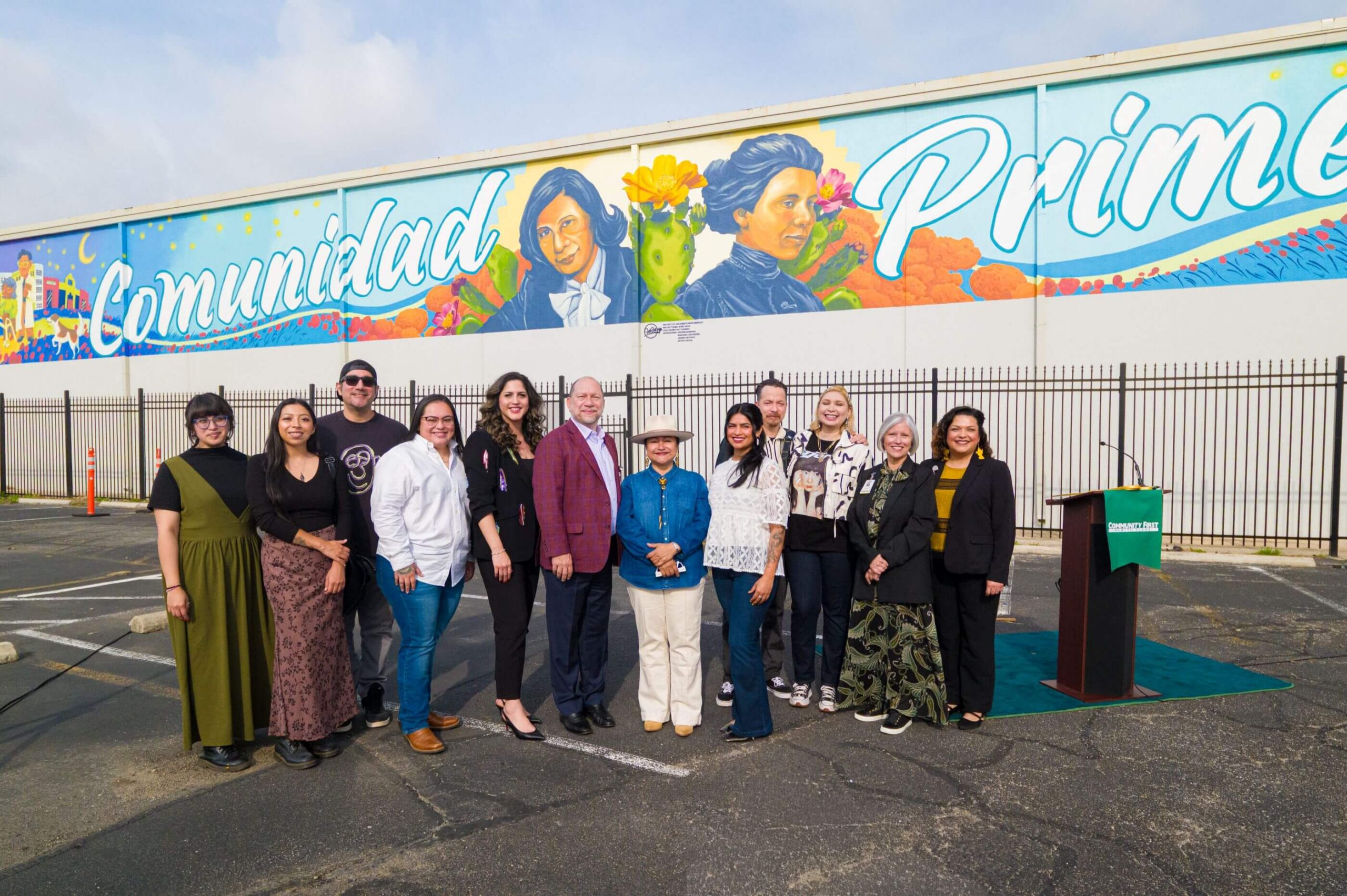 A group of people stands in front of a large, colorful outdoor mural featuring the word "Comunidad" and portraits of historical figures.