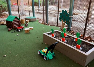 A brightly colored outdoor play area featuring a toy log cabin, a red wagon, wooden stumps, a toy lawnmower, and a raised garden bed with toy carrots, enclosed by wire fencing.