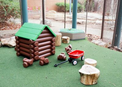 A brown plastic log cabin with a green roof sits on a green artificial turf playground next to a red Radio Flyer wagon and wooden log stools, enclosed by a chain-link fence.