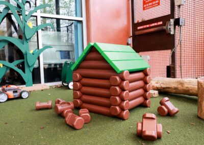 Plastic toy log cabin with a green roof sits on green turf near cutout corn stalks and an emergency exit door.