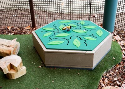 Outdoor sensory play station with a hexagonal top featuring a bee figure, green leaves, and black vine designs, situated on artificial turf near wooden logs and leaf litter.