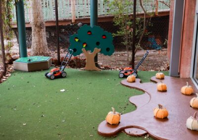 An indoor play area features artificial turf, two toy lawnmowers, a cutout apple tree, and several fabric pumpkins resting on a brown, winding play surface.