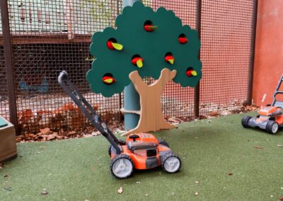Two toy lawnmowers sit on artificial turf next to a wooden play apple tree structure with red and yellow apples.