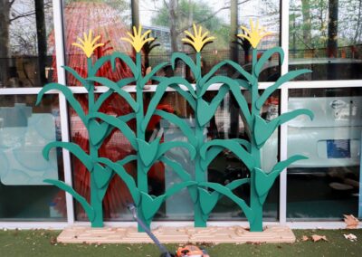 Decorative, stylized wooden cutouts of green corn stalks with yellow tassels are displayed in front of a large window reflecting an outdoor scene.