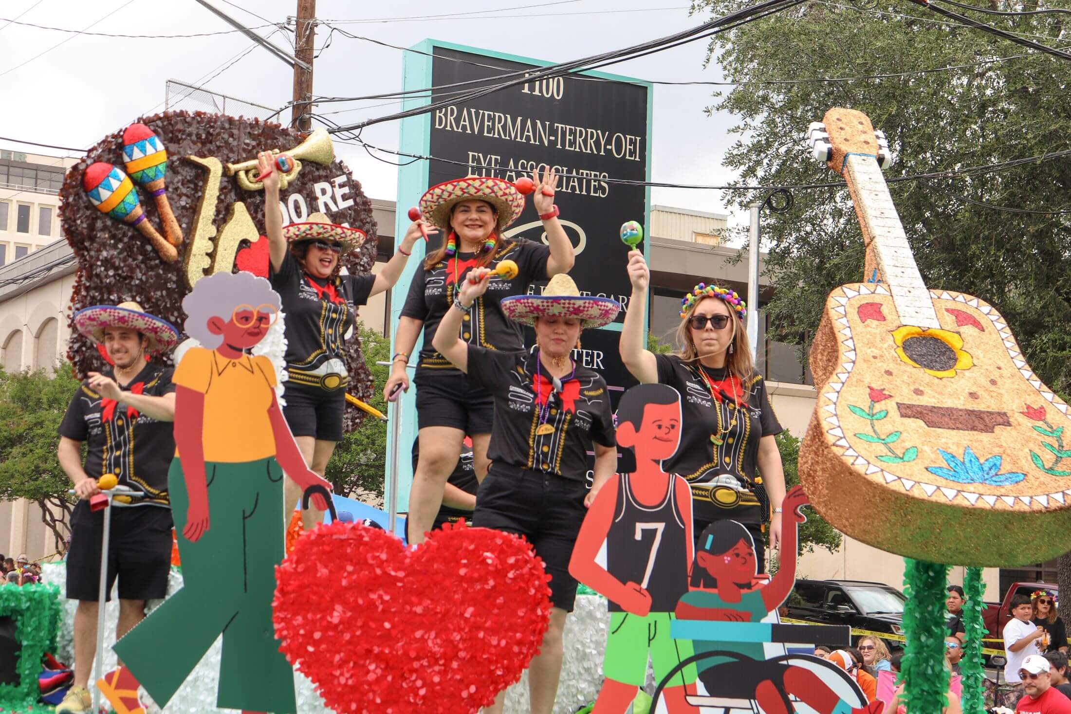People in sombreros celebrating on a parade float decorated with a giant guitar, maracas, and cutout figures against a backdrop of a business sign.