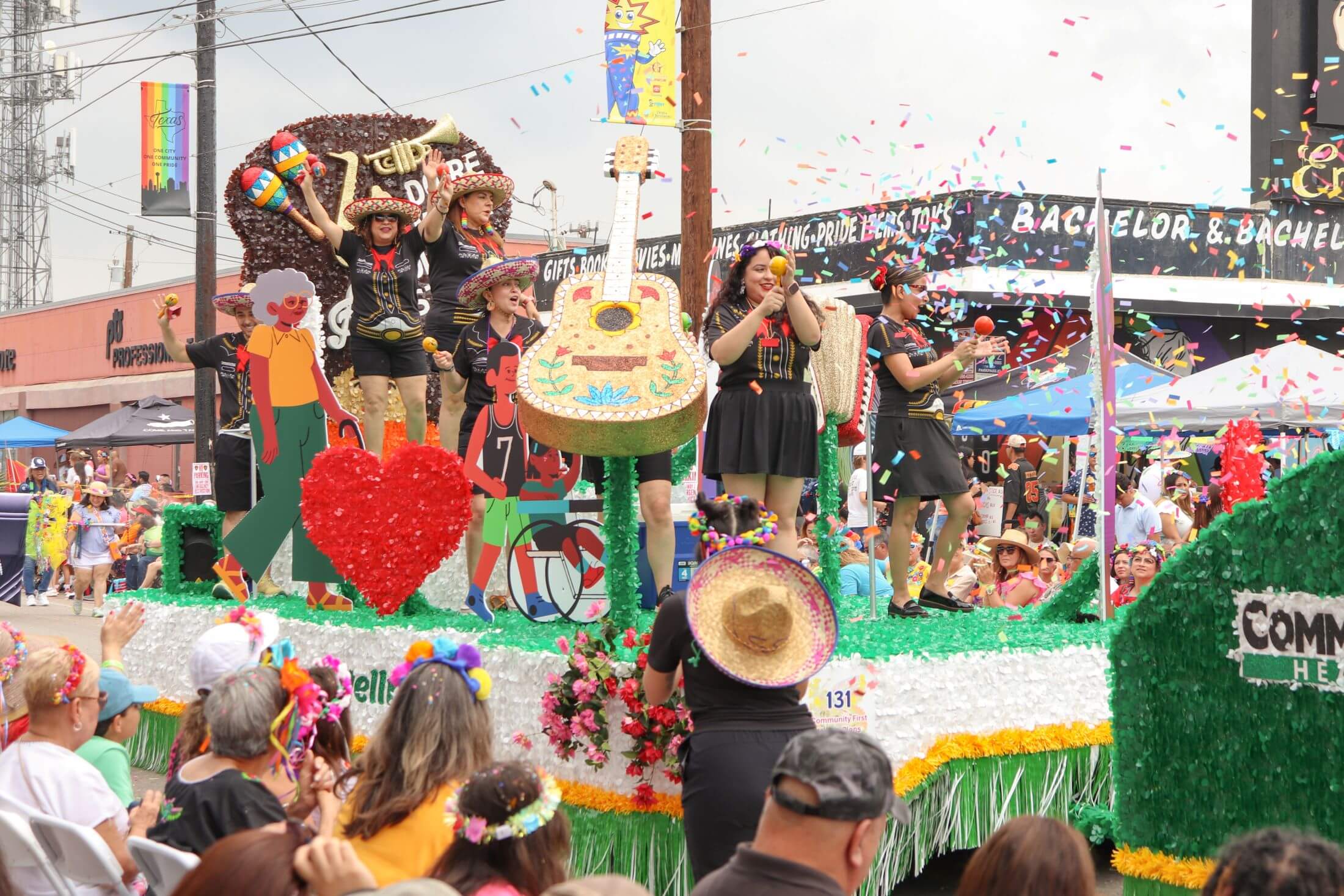 A colorful parade float decorated with a large guitar and mariachi-attired performers tosses confetti over a crowd during a daytime festival.