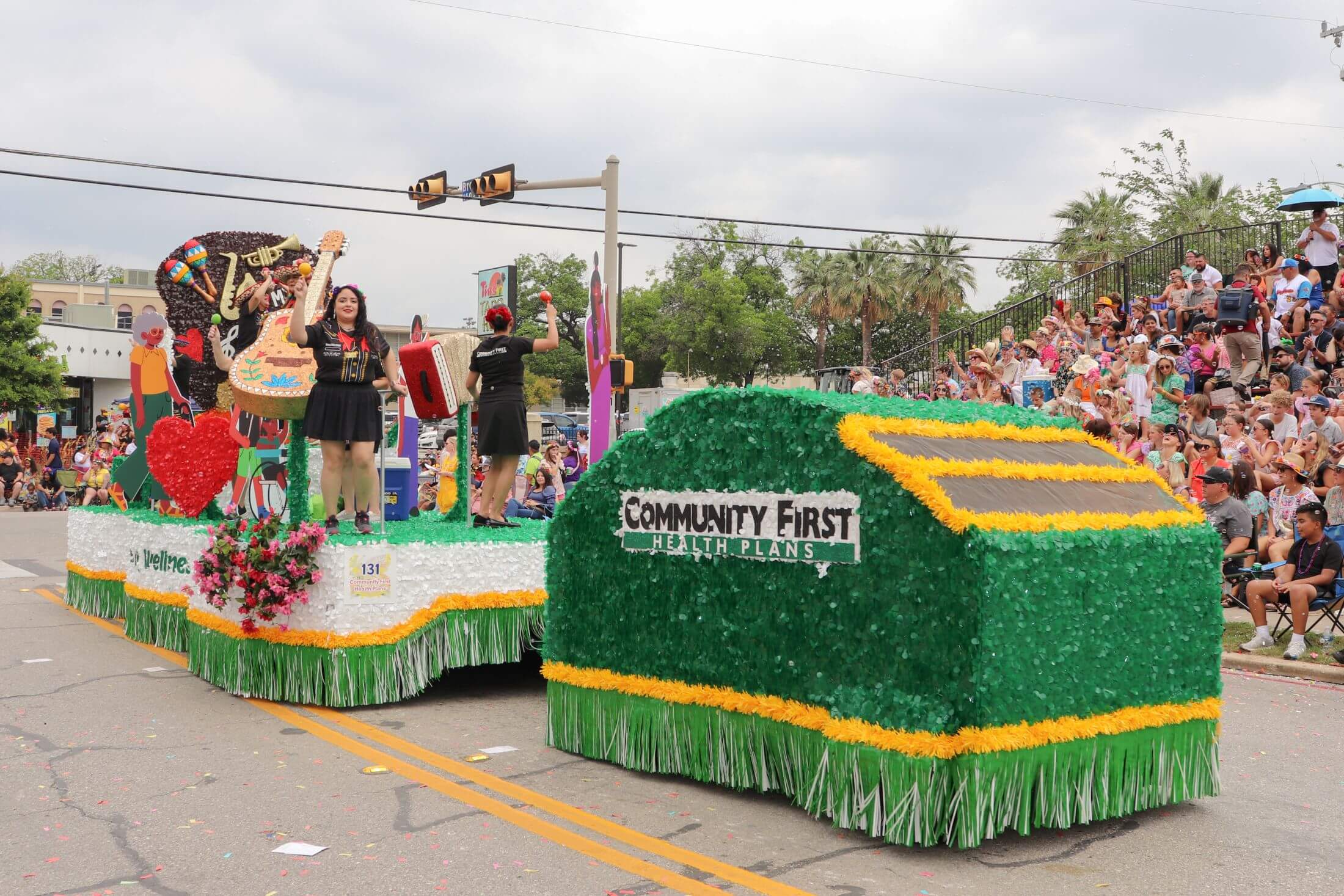 A vibrant parade float featuring volunteers with musical instruments and a large green "Community First Health Plans" section drives past a large crowd seated along the street.